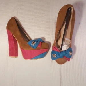 Färgglada pumps med rosett - Unika klackar i brunt, rosa och blått,med rosett med döskalle dekorationer i metall. Hög klack och stabil platåsula som ger en bra bekväm vinkel för foten. 