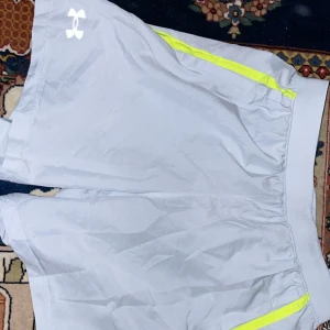Grå Springshorts från Under Armour - Snygga grå träningsshorts från Under Armour med neongula detaljer. Perfekta för gymmet eller löprundan. De har en bekväm passform och är tillverkade i ett lätt och andningsbart material.