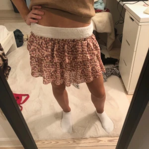 Rosa blommig kortkjol - Söt rosa kortkjol med blommigt mönster och volangdetaljer. Kjolen har en bred vit resår i midjan för bekväm passform. Perfekt för varma dagar och ger en lekfull look.