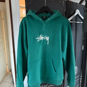 Grön hoodie från Stussy - Säljer en snygg grön hoodie från Stussy med deras ikoniska logga på bröstet. Tröjan har en klassisk känguruficka och justerbar huva med dragsko. Perfekt för en avslappnad stil.