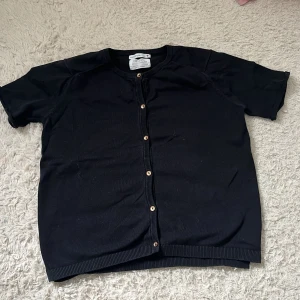 Svart kofta med knappar - Säljer en klassisk svart kofta med knappar framtill. Den har korta ärmar och ribbade kanter vid ärmslut och nederkant. Perfekt för lager-på-lager-stil eller som ett lättare ytterplagg.