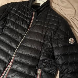 Moncler Daniel - Säljer en snygg svart dunjacka från Moncler med quiltad design och dragkedja. Jackan har handgjord Moncler-logga på ärmen och en stilren look. Perfekt för kyliga dagar! Pyttelitet hål på högerärmen (läcker ej dun)