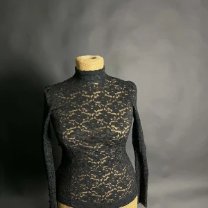Svart spetsblus med knappar - Elegant svart spetsblus med långa ärmar och knappar framtill. Blusen har en hög krage och ett vackert spetsmönster som ger en sofistikerad look. Perfekt för att lyfta vilken outfit som helst.