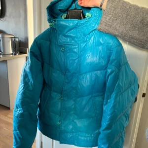 Blå dunjacka från WeSC - Säljer en blå dunjacka från WeSC med huva och dragkedja. Jackan har en pufferdesign och är perfekt för kyligare väder. Den har en stilren look med en glansig yta och justerbara detaljer vid huvan.