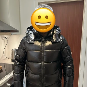Svart dunjacka från Moncler - Säljer en snygg svart dunjacka från Moncler. Jackan har en glansig finish och är utrustad med en dragkedja framtill. Den har en stor huva och långa ärmar, perfekt för kyliga dagar. Moncler-loggan syns på ärmen. Perfekt för vintern!