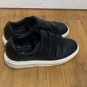 Svarta sneakers med kardborreband från Zara - Snygga svarta sneakers från Zara med praktiska kardborreband. Skorna har en vit sula som ger en fräsch kontrast. Perfekta för en stilren look finns ingen skador absolut men det är använda inte så mycket men den är som ny 