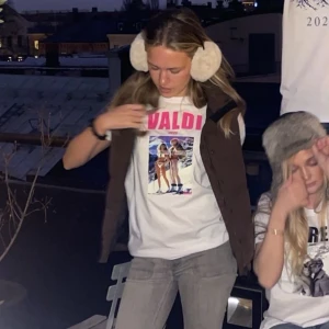 Vit t-shirt med Valdi-tryck - Vit t-shirt med färgglatt Valdi-tryck på framsidan. T-shirten har en klassisk passform och korta ärmar, perfekt för en avslappnad stil. Trycket visar en retroinspirerad skidscen med texten 'Valdi' i rött alternativt rosa ovanför.