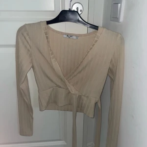 Beige omlottopp från NA-KD - Snygg beige omlottopp från NA-KD i ribbat material. Toppen har långa ärmar och en v-ringad design med knytband i midjan. Perfekt för en stilren look.