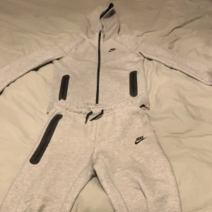 Grå hoodie och byxor från Nike tech fleece - Säljer en grå hoodie och matchande byxor från Nike. Hoodien har dragkedja och långa ärmar, medan byxorna har en bekväm resår i midjan. Perfekt för en avslappnad stil.