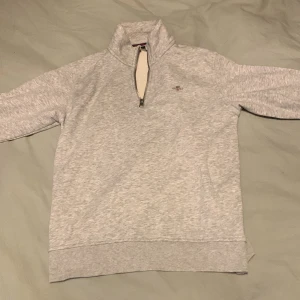 Grå tröja med dragkedja från Gant - Säljer en stilren grå tröja från Gant med halv dragkedja och hög krage. Perfekt för en avslappnad look. Tröjan har långa ärmar och ribbade muddar vid ärmslut och nederkant. Passar bra till både jeans och chinos.