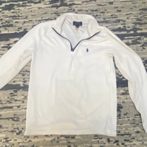 Vit långärmad tröja från Polo Ralph Lauren - Säljer en stilren vit långärmad tröja från Polo Ralph Lauren med en klassisk krage och dragkedja. Tröjan har det ikoniska broderade logotypen på bröstet. Perfekt för en avslappnad och elegant look.