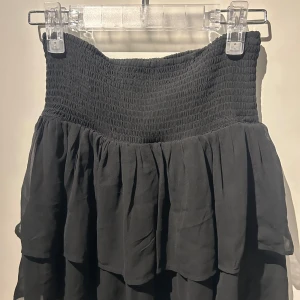 Svart kjol från KappAhl - Snygg svart kjol från KappAhl med smockdetaljer i midjan och volanger. Perfekt för en stilren look. Kjolen är kort och har en luftig passform.