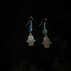 Silverfärgade örhängen med hamsa och öga - Hand gjorda silverfärgade örhängen med hamsa-händer och blå ögon. Perfekta för att ge en mystisk touch till din stil. Hängena är detaljerade och har en krok för enkel användning. 