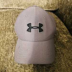 Grå keps från Under Armour - Snygg grå keps från Under Armour med broderad logga framtill. Perfekt för sportiga aktiviteter eller som en stilren accessoar. Kepsen har en klassisk design med böjd skärm och justerbar passform.