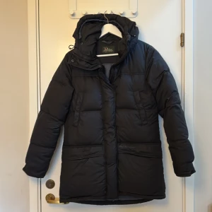 Svart dunjacka från Peak Performance - Fluffig och härlig svart dunjacka med luva ifrån Peak performance. Modell Snowline J.   4 fickor på utsidan. Varav 2 fickor med fleecefoder och dragkedja och 2 högre upp med knapp. 2 fickor inuti jackan, varav 1 med dragkedja.