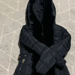 Svart dunjacka med pälsdetaljer - Snygg svart dunjacka med huva och mjuka pälsdetaljer längs med kragen. Jackan har en dragkedja framtill och är perfekt för kyliga dagar. Den quiltade designen ger en stilren look.