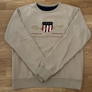 Beige sweatshirt från GANT - Säljer en stilren beige sweatshirt från GANT med broderad logotyp och text på framsidan. Tröjan har långa ärmar och ribbade muddar vid ärmslut och nederkant. Perfekt för en avslappnad stil.