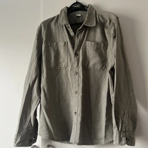 Grön skjorta från Lindex - Säljer en snygg grön skjorta från Lindex i storlek 116. Skjortan har långa ärmar och två bröstfickor med knappar. Perfekt för både vardag och lite finare tillfällen. Materialet känns mjukt och bekvämt, vilket gör den skön att bära hela dagen. Passar bra till både jeans och chinos!