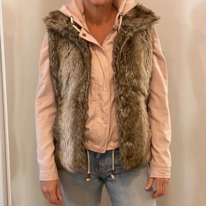 Brun pälsväst från Livelo - Säljer en snygg brun pälsväst från Livelo, perfekt för höst och vinter. Västen har ett bälte i midjan för en tight passform och är i bra skick. Den är designad i Sverige och ger en lyxig känsla till vilken outfit som helst.