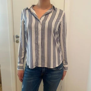 Randig skjorta i blått och vitt - Säljer en stilren randig skjorta i blått och vitt. Skjortan har långa ärmar och en klassisk krage. Perfekt för både vardag och festliga tillfällen. Materialet känns mjukt och bekvämt mot huden. Passar bra till jeans för en avslappnad look. Uppskattar storlek M