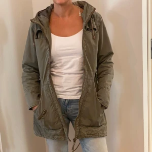 Olivgrön parkajacka med huva - Säljer en olivgrön parkajacka i bra skick. Jackan har en praktisk huva och dragkedja framtill. Perfekt för höst och vår med sina långa ärmar och bekväma passform. Den har också flera fickor för extra förvaring. Perfekt för vardagsbruk!