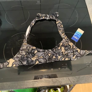 Svart och beige blommig bikiniöverdel från Uniq - Snygg bikiniöverdel i svart och beige med blommigt mönster. Den har en knytning i nacken och en bekväm passform. Perfekt för sommarens stranddagar! 🌸