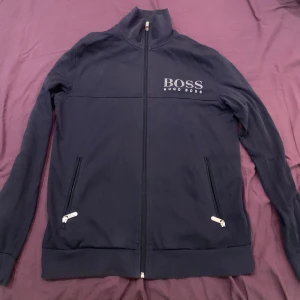 Hugo boss tröja - Säljer en stilren svart kofta från Hugo Boss. Den har en dragkedja framtill och två praktiska fickor med dragkedjor. Perfekt för höst och vår, med en normal passform och långärmade ärmar. Tröjan är i bra skick och redo för nya äventyr!