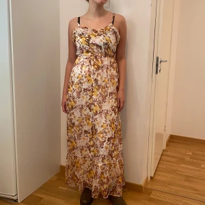 Blommig långklänning med volangdetaljer - Säljer en superfin blommig långklänning i storlek 34. Klänningen har ett vackert mönster i gult och brunt med volangdetaljer. Perfekt för sommarens fester eller en mysig dag i solen. Den är ärmlös och har en luftig passform som gör den bekväm att bära hela dagen.