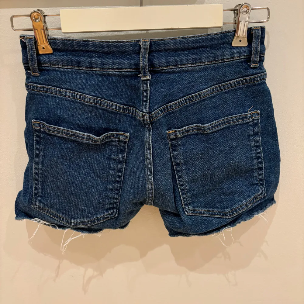 Säljer ett par snygga blå lågmidjadd jeansshorts med fransiga kanter. De har en klassisk femficksdesign och knappgylf. Perfekta för sommardagar och festivaler! 🕶️. Shorts.