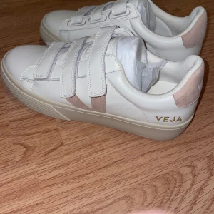 Vita sneakers från Veja - Snygga vita sneakers från Veja med tre praktiska kardborreband. Skorna har en stilren design med ljusrosa detaljer på hälen och sidan. Perfekta för en avslappnad och trendig look. Sulan är lite gul från design, de har aldrig burits.