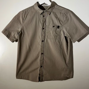 Beige kortärmad skjorta från H&M - Säljer en stilren beige kortärmad skjorta från H&M. Skjortan har en klassisk krage och knappar framtill samt en bröstficka med knapp. Perfekt för en avslappnad look.