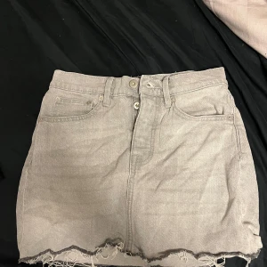 Grå jeanskjol - Säljer en cool grå jeanskjol med rå kant och klassisk femficksdesign. Kjolen har en knapp- och dragkedjestängning framtill och bälteshällor. Perfekt för en avslappnad stil.