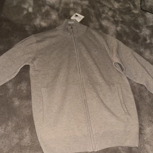 Beige ulltröja från GANT - Säljer en stilren beige tröja från GANT i superfin lammull. Tröjan har en dragkedja framtill och ribbade detaljer vid kragen. Perfekt för kyligare dagar.