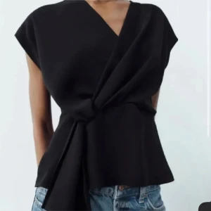 Svart omlottopp ZARA - Snygg svart omlottopp med korta ärmar och en elegant drapering framtill. Perfekt för en stilren look, passar bra till jeans eller kjol.