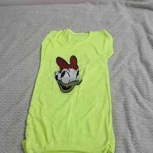 Gul t-shirtklänning med tryck - Säljer en cool gul t-shirtklänning med ett tryck av en tecknad figur på framsidan. Klänningen har en avslappnad passform och är perfekt för en avslappnad stil. Den är ärmlös och har en rund halsringning.