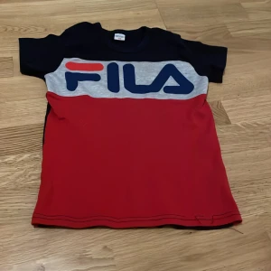 Svart och röd t-shirt från FILA - Säljer en cool t-shirt från FILA i svart och rött med stor logga på framsidan. T-shirten är kortärmad och perfekt för en avslappnad stil. Tillverkad i Turkiet.