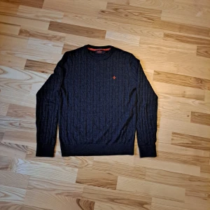 Mörkgrå tröja från Morris - Säljer en stilren mörkgrå tröja från Morris med kabelstickat mönster. Tröjan har en liten broderad logga på bröstet och rund halsringning. Perfekt för kyligare dagar.