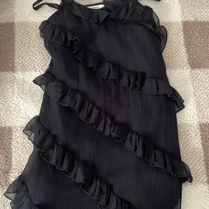 Svart klänning med volanger från Gina Tricot - Snygg svart klänning från Gina Tricot med volanger som går diagonalt över hela klänningen. Den har justerbara axelband och en luftig passform. Perfekt för en stilren look!