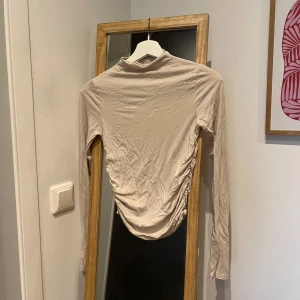 Beige långärmad topp - Säljer en stilren beige långärmad topp med rynkade detaljer på sidorna. Toppen har en hög krage och är tillverkad i ett mjukt material. Den är i strl S och är från Gina Tricot. Sparsamt använd och är som ny✨