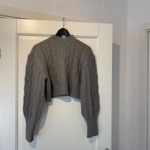 Grå stickad croppad tröja - Säljer en snygg grå stickad tröja med kabelstickat mönster. Tröjan är croppad och har långa ärmar med ribbad krage. Perfekt för en trendig look!