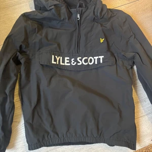 Svart vindjacka från Lyle & Scott - Säljer en svart vindjacka från Lyle & Scott Junior. Jackan har en stor ficka framtill med dragkedja och logotyp, samt en praktisk huva. Perfekt för blåsiga dagar och har en stilren design.
