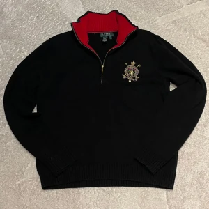 Svart tröja med broderad logga från Ralph Lauren - Säljer en svart tröja från Ralph Lauren med en snygg broderad logga på bröstet. Tröjan har en dragkedja vid halsen och en röd insida på kragen som ger en fin kontrast. Perfekt för en stilren look. Det står L men passar 14-16