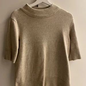 Beige stickad tröja - Säljer en stilren beige stickad tröja med korta ärmar och ribbade detaljer vid hals, ärmslut och nederkant. Perfekt för en avslappnad look. Passar bra till både jeans och kjol.