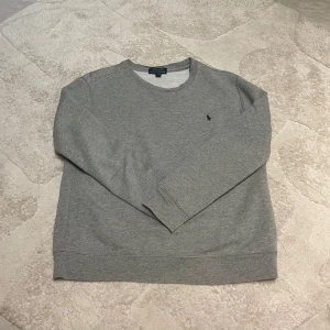 Grå sweatshirt från Polo Ralph Lauren - Säljer en stilren grå sweatshirt från Polo Ralph Lauren med det klassiska logotypbroderiet på bröstet. Tröjan har långa ärmar och en rund halsringning, perfekt för en avslappnad look. Passar bra till både jeans och chinos. 15-16