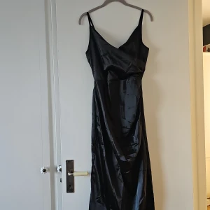 Svart långklänning i polyester - Elegant svart långklänning med smala axelband och omlott-design. Tillverkad i glansig polyester som ger en lyxig känsla. Perfekt för en stilren look.