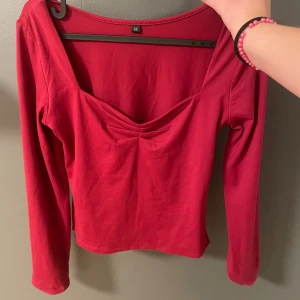 Röd långärmad topp - Säljer en snygg röd långärmad topp med v-ringning och rynkdetalj framtill. Toppen har en enkel design och är perfekt för en stilren look. Materialet känns mjukt och bekvämt mot huden.