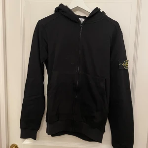 Svart Stone Island Zip Hoodie - Säljer en svart hoodie från Stone Island med dragkedja och huva. Den har en ikonisk logga på ärmen och är perfekt för en stilren look. Passar bra till både vardag och avslappnade tillfällen. Skick 8/10 saknar snören.