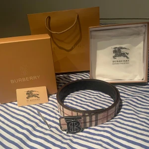 Burberry bälte - Säljer ett stilrent bälte från Burberry med klassiskt rutigt mönster i beige, svart och rött. Bältet har en elegant spänne med Burberry-logotypen. Perfekt för att ge en touch av lyx till din outfit. Man kan byta sida på bältet så att du har svart ist för beige!