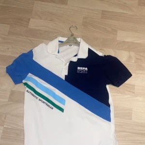 Vit och blå pikétröja från USPA Sport - Snygg vit pikétröja med blå och gröna detaljer från USPA Sport. Tröjan har en klassisk krage och korta ärmar. Perfekt för en sportig look med texten 'Authentic Sportswear' på framsidan.