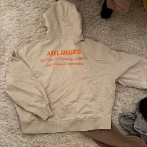 Beige hoodie från Axel Arigato - ALDRIG ANVÄND Säljer en beige hoodie från Axel Arigato med tryck i orange på både fram- och baksidan. Hoodien har en klassisk design med en stor ficka framtill och ribbade muddar vid ärmslut och nederkant. Perfekt för en avslappnad stil.
